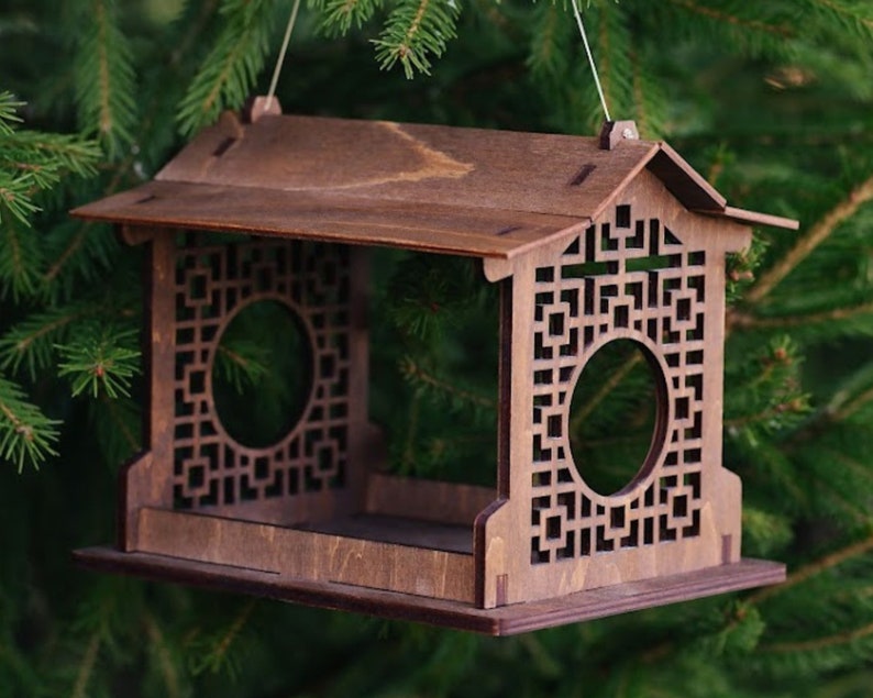 Op de afbeelding: Een houten vogelvoederhuisje met een decoratief, geometrisch ontwerp. De voederbak heeft een dak en een platform waar vogels op kunnen zitten. Het hangt aan een tak.