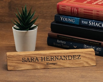 May include: A wooden desk nameplate with the name "SARA HERNANDEZ" and the title "Manager" engraved in black. A small potted succulent and a stack of books are in the background. The nameplate is made of light brown wood.