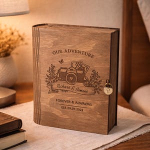 Könnte beinhalten: Eine holzfarbene, buchförmige Schatulle mit Schloss, graviert mit den Worten "OUR ADVENTURE" und "Richard & Simone". Das Design umfasst eine Kamera und florale Elemente. Auf der Unterseite steht "FOREVER & ALWAY.NS EST. 09.07.2023."