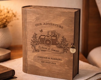May include: A wooden book-shaped box with a lock, engraved with the words "OUR ADVENTURE" and "Richard & Simone." The design includes a camera and floral elements. The bottom of the box reads "FOREVER & ALWAY.NS EST. 09.07.2023."