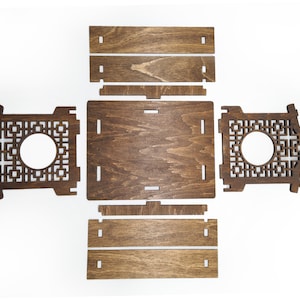 Op de afbeelding: Een houten doos met een decoratief uitgesneden ontwerp. De doos is gemaakt van donkerbruin hout en heeft een vierkante vorm. De doos is gedemonteerd en plat gelegd, waardoor de afzonderlijke onderdelen zichtbaar zijn.