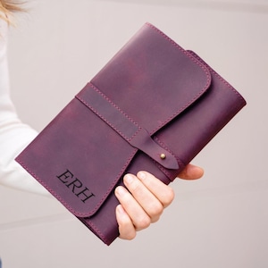 May include: A deep purple leather journal cover featuring a wrap-around strap with a brass stud closure. The front flap is embossed with the letters ERH in a serif font. The cover has a smooth matte texture and visible stitching along the edges.