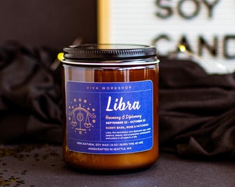May include: A brown glass candle jar with a black lid. The label is blue with gold lettering and features the zodiac sign Libra. The text on the label reads "Libra Harmony & Diplomacy September 23 - October 22 Scent: Basil, Rose & Sandalwood 100% Natural Soy Wax | 8 oz | Handcrafted in Seattle, WA"