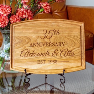 May include: A wooden cutting board with a curved top edge, engraved with "35th Anniversary Aleksandr & Alla EST. 1988". The board is displayed on a black metal stand.