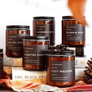 May include: An assortment of glass jar candles with black lids and labels. The labels display text such as "Cranberry Prosecco," "Oakmoss & Amber," "Fallen Leaf," "Morning Coffee," "Pumpkin Spice," "Campfire Nights," and "Cozy Weather." The candles are arranged on books and a patterned cloth.