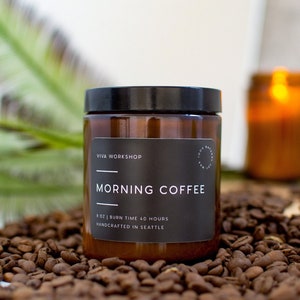 May include: A brown glass candle jar with a black label that reads "Viva Workshop Morning Coffee 4 oz | Burn Time 40 Hours Handcrafted in Seattle". The candle is sitting on a bed of coffee beans.