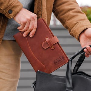 May include: A rectangular brown leather notebook with a strap closure and a small embossed label reading KHG, being placed into a black and gray duffel bag. The notebook features visible stitching along the edges and a smooth, matte finish.