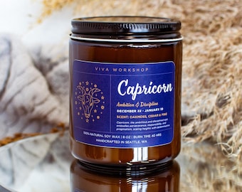 May include: A brown glass candle jar with a blue label that says "Viva Workshop" and "Capricorn". The label also includes the dates "December 22 - January 19" and the text "Ambition & Discipline". The scent is described as "Dark, Sandalwood, Cedar & Pine".