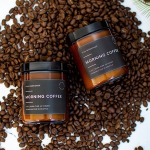 May include: Two amber glass candle jars with black lids and labels. The labels read "Viva Workshop" and "Morning Coffee". The candles are surrounded by coffee beans.