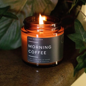 May include: A brown glass candle jar with a black label that reads "Viva Workshop Morning Coffee 4 oz | Burn Time 20 Hours Handcrafted in Seattle". The candle is lit and the flame is visible.