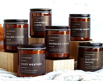 May include: An assortment of glass jar candles with black lids and labels. The labels feature text such as "Pumpkin Spice," "Fallen Leaves," and "Campfire Nights." The candles are arranged on wooden blocks and a white surface, with fall leaves in the background.