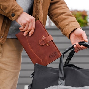 May include: A brown leather journal with a buckle closure and the initials "KHG" engraved on the front. The journal is held next to a black and gray duffel bag. The person is wearing a brown jacket and gray shirt.