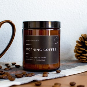 May include: A brown glass candle jar with a black label that reads "Viva Workshop Morning Coffee 8 oz | Burn Time 40 Hours Handcrafted in Seattle". The candle is sitting on a white and brown plaid fabric with coffee beans scattered around it.
