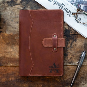 Peut inclure: Un journal en cuir marron avec une fermeture &agrave; lani&egrave;re et un motif d&eacute;coratif. Le journal est pos&eacute; sur une surface en bois, &agrave; c&ocirc;t&eacute; d'une tasse de caf&eacute;, d'un stylo et d'un journal.