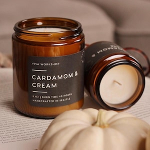 Puede incluir: Dos velas en frasco de vidrio &aacute;mbar con etiquetas negras. La etiqueta frontal dice VIVA WORKSHOP CARDAMOM & CREAM 8 OZ | BURN TIME 40 HOURS HANDCRAFTED IN SEATTLE. Las velas contienen cera blanca y una mecha, junto a peque&ntilde;as calabazas blancas.