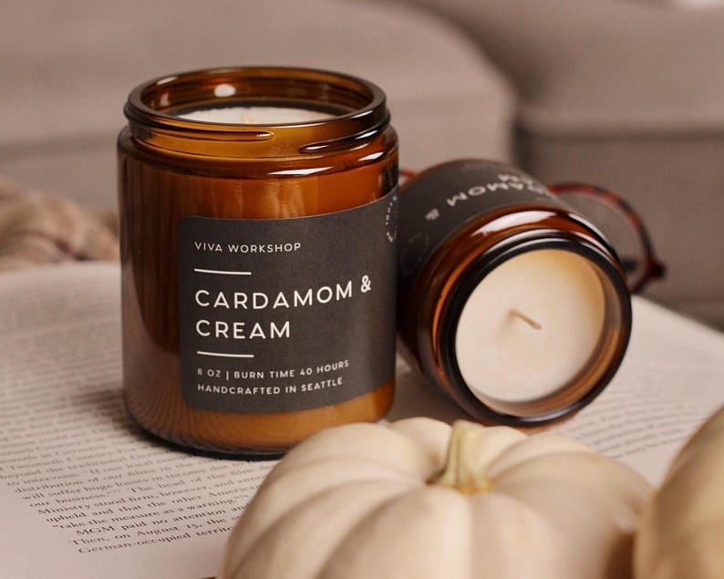 Puede incluir: Dos velas en frasco de vidrio &aacute;mbar con etiquetas negras. La etiqueta frontal dice VIVA WORKSHOP CARDAMOM & CREAM 8 OZ | BURN TIME 40 HOURS HANDCRAFTED IN SEATTLE. Las velas contienen cera blanca y una mecha, junto a peque&ntilde;as calabazas blancas.