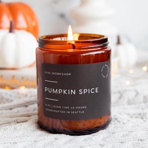 Puede incluir: Una vela encendida en un frasco de vidrio &aacute;mbar con una etiqueta negra que dice VIVA WORKSHOP PUMPKIN SPICE 8 OZ | BURN TIME 40 HOURS HANDCRAFTED IN SEATTLE. La vela tiene una sola llama y est&aacute; colocada frente a calabazas naranjas.