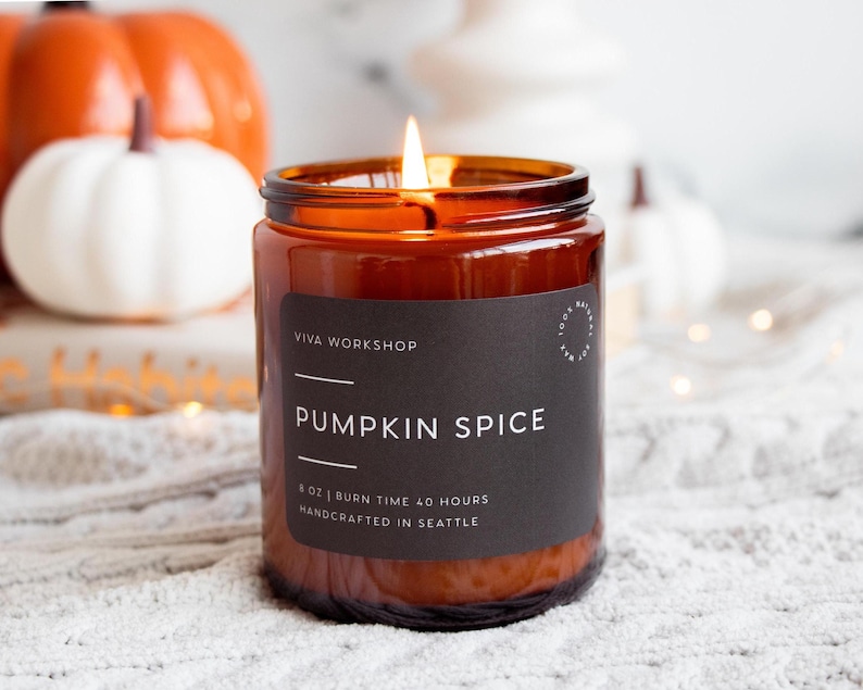 Puede incluir: Una vela encendida en un frasco de vidrio &aacute;mbar con una etiqueta negra que dice VIVA WORKSHOP PUMPKIN SPICE 8 OZ | BURN TIME 40 HOURS HANDCRAFTED IN SEATTLE. La vela tiene una sola llama y est&aacute; colocada frente a calabazas naranjas.