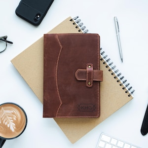 Peut inclure: Un journal en cuir marron avec un fermoir, pos&eacute; sur un carnet &agrave; spirales. Un stylo argent&eacute;, des lunettes, une montre avec un bracelet noir, un t&eacute;l&eacute;phone, une tasse de caf&eacute; et un clavier sont &eacute;galement sur la surface blanche.