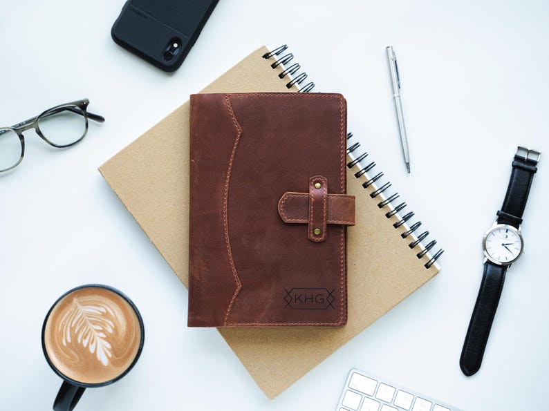 Peut inclure: Un journal en cuir marron avec un fermoir, pos&eacute; sur un carnet &agrave; spirales. Un stylo argent&eacute;, des lunettes, une montre avec un bracelet noir, un t&eacute;l&eacute;phone, une tasse de caf&eacute; et un clavier sont &eacute;galement sur la surface blanche.