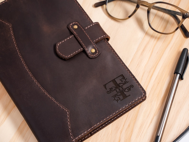 Peut inclure: Un journal en cuir marron fonc&eacute; avec une initiale d&eacute;corative et le nom "LEE" grav&eacute; sur la couverture. Le journal a une fermeture &agrave; lani&egrave;re avec des rivets en laiton. Un stylo et des lunettes sont &eacute;galement visibles.