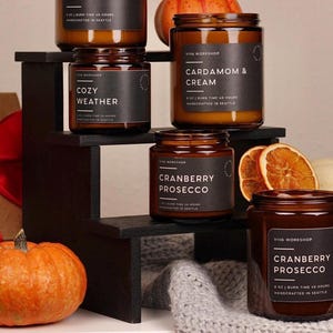 Puede incluir: Cinco velas en frascos de vidrio &aacute;mbar con etiquetas negras y texto blanco que incluye Viva Workshop, Cozy Weather, Cardamom & Cream y Cranberry Prosecco. Las velas est&aacute;n dispuestas en un soporte negro escalonado con calabazas y rodajas de naranja seca.