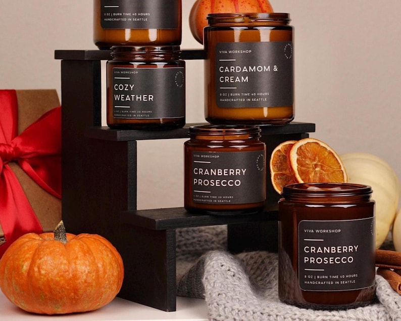 Puede incluir: Cinco velas en frascos de vidrio &aacute;mbar con etiquetas negras y texto blanco que incluye Viva Workshop, Cozy Weather, Cardamom & Cream y Cranberry Prosecco. Las velas est&aacute;n dispuestas en un soporte negro escalonado con calabazas y rodajas de naranja seca.