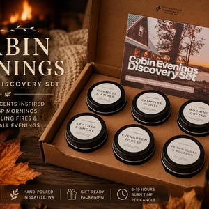 May include: A cardboard box containing six small black metal tins of candles, each with a white label. The labels read Oakmoss & Amber, Campfire Nights, Morning Coffee, Leather & Smoke, Evergreen Forest, and Brown Sugar & Bourbon. A card titled Cabin Evenings Discovery Set is included.