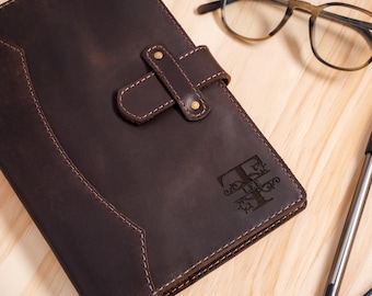 May include: A dark brown leather journal cover featuring a secure strap with two brass-colored metal rivets. The cover is embossed with a decorative letter T and the name LEE. The leather has visible stitching along the edges and a smooth texture.