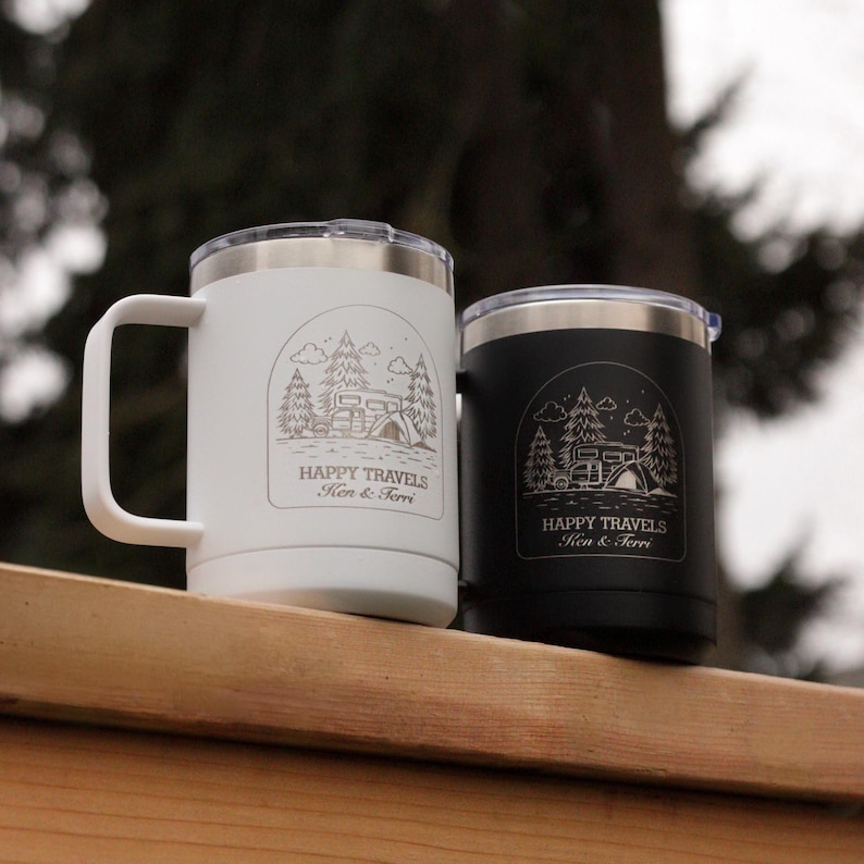 Puede incluir: Dos tazas de viaje aisladas, una blanca y otra negra, con tapas y asas plateadas. Cada taza presenta un gr&aacute;fico con tem&aacute;tica de camping con el texto "HAPPY TRAVELS Ken & Terri."