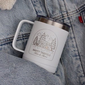 Puede incluir: Taza de viaje blanca con borde y asa plateados. La taza presenta un gr&aacute;fico con tem&aacute;tica de camping con &aacute;rboles, una caravana y las palabras "HAPPY TRAVELS" y "Ken & Terri" en un marco rectangular.