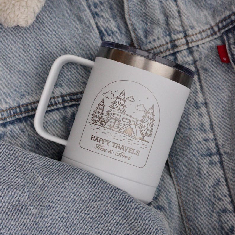 Puede incluir: Taza de viaje blanca con borde y asa plateados. La taza presenta un gr&aacute;fico con tem&aacute;tica de camping con &aacute;rboles, una caravana y las palabras "HAPPY TRAVELS" y "Ken & Terri" en un marco rectangular.