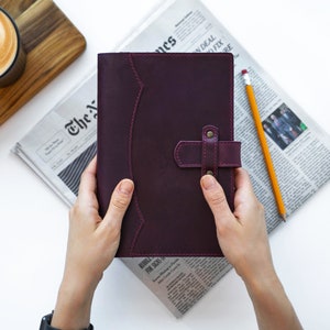 Peut inclure: Un journal en cuir violet fonc&eacute; avec une fermeture &agrave; lani&egrave;re, tenu par des mains. Le journal est pos&eacute; sur un journal avec un crayon et une petite plante succulente &agrave; proximit&eacute;. Une tasse de caf&eacute; avec du latte art est en arri&egrave;re-plan.
