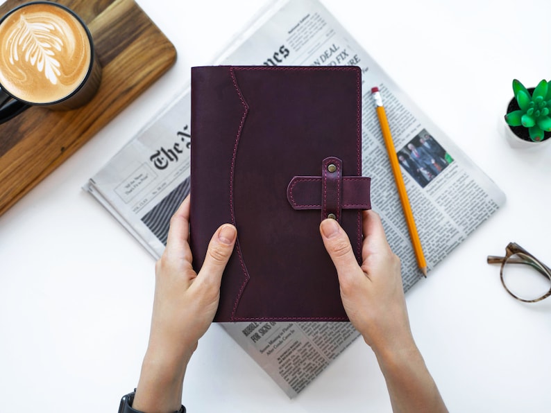 Peut inclure: Un journal en cuir violet fonc&eacute; avec une fermeture &agrave; lani&egrave;re, tenu par des mains. Le journal est pos&eacute; sur un journal avec un crayon et une petite plante succulente &agrave; proximit&eacute;. Une tasse de caf&eacute; avec du latte art est en arri&egrave;re-plan.