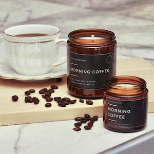 Puede incluir: Dos velas en frasco de vidrio &aacute;mbar con etiquetas negras Morning Coffee, una taza de t&eacute; blanca sobre un platillo y granos de caf&eacute; tostado esparcidos. La vela grande indica 255g y 45 horas, la vela peque&ntilde;a indica 113g de peso.