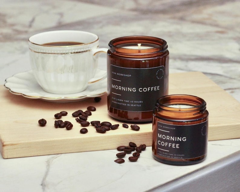 Puede incluir: Dos velas en frasco de vidrio &aacute;mbar con etiquetas negras Morning Coffee, una taza de t&eacute; blanca sobre un platillo y granos de caf&eacute; tostado esparcidos. La vela grande indica 255g y 45 horas, la vela peque&ntilde;a indica 113g de peso.