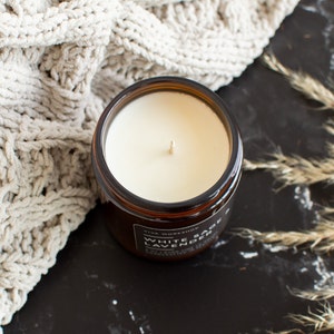 May include: A brown glass candle jar with a white soy wax candle inside. The candle has a single wick and the jar has a black label that reads "White Sage & Lavender" with the words "Viva Workshop" above the scent name.