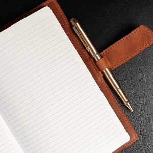 Peut inclure: Un carnet ouvert avec des pages lign&eacute;es, reli&eacute; en cuir marron. Un stylo de couleur or est maintenu dans une boucle en cuir sur le c&ocirc;t&eacute; droit. Le carnet est pos&eacute; sur une surface noire.