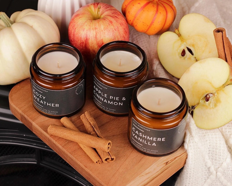 Puede incluir: Tres velas en frascos de vidrio &aacute;mbar con etiquetas negras que dicen Cozy Weather, Apple Pie & Cinnamon y Cashmere Vanilla. Las velas est&aacute;n dispuestas con calabazas blancas y naranjas, manzanas enteras y en rodajas, y ramas de canela.