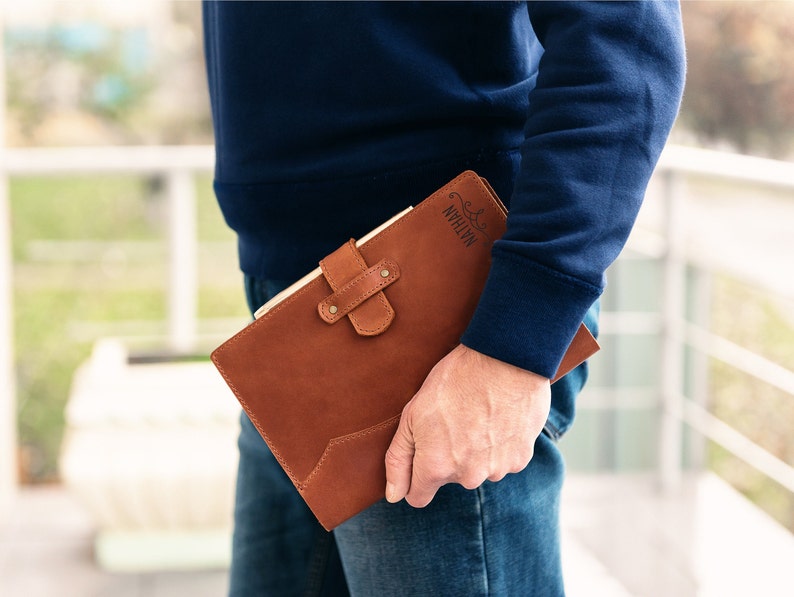 May include: A brown leather journal with the name 'Nathan' engraved on the cover. The journal has a clasp closure and a strap.  It features a minimalist design and is shown held in a person's hand.