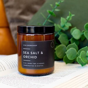 Puede incluir: Un tarro de vidrio marr&oacute;n con una vela, una tapa negra y una etiqueta. La etiqueta dice "Viva Workshop", "Sea Salt & Orchid", "8 oz | Burn Time 40 Hours", y "Handcrafted in Seattle".