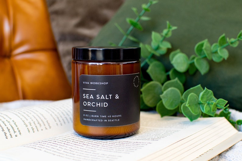 Puede incluir: Un tarro de vidrio marr&oacute;n con una vela, una tapa negra y una etiqueta. La etiqueta dice "Viva Workshop", "Sea Salt & Orchid", "8 oz | Burn Time 40 Hours", y "Handcrafted in Seattle".