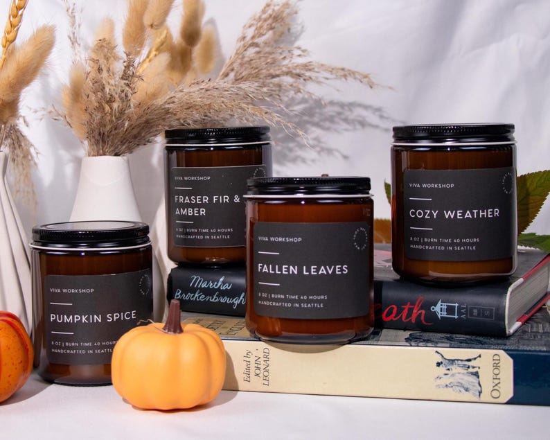 Puede incluir: Cuatro velas en frasco de vidrio &aacute;mbar con tapas y etiquetas negras. Las etiquetas dicen Viva Workshop, 9 oz, tiempo de combusti&oacute;n 40 horas, hecho a mano en Seattle, y los aromas Pumpkin Spice, Fraser Fir & Amber, Fallen Leaves, Cozy Weather.
