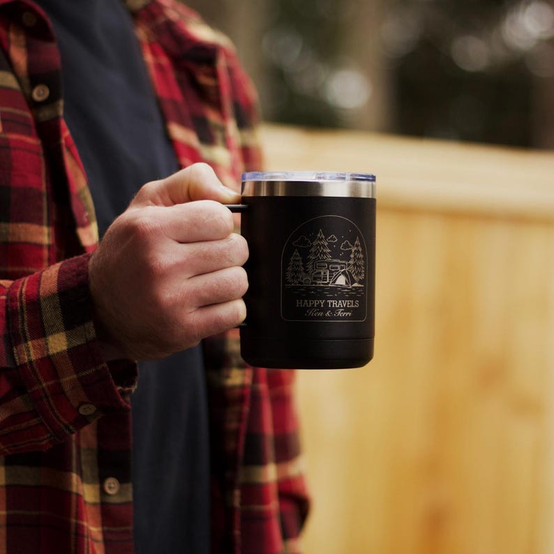Puede incluir: Taza de viaje negra con borde y asa plateados. La taza presenta una escena de acampada con &aacute;rboles, una caravana y las palabras "HAPPY TRAVELS" y un nombre. La taza la sostiene una persona que lleva una camisa a cuadros roja y negra.