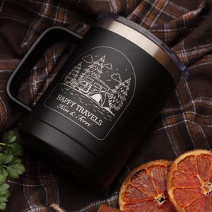 Puede incluir: Taza de viaje negra con asa y borde plateado. La taza presenta un gr&aacute;fico blanco de una caravana, &aacute;rboles y una tienda de campa&ntilde;a, con las palabras "Happy Travels" y un nombre debajo. La taza est&aacute; sobre una superficie a cuadros.