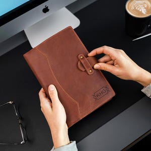 Peut inclure: Un journal en cuir marron avec une fermeture &agrave; boucle. Le journal pr&eacute;sente un motif cousu et les initiales "KHG" sont visibles. Le journal est tenu par une personne. Une tasse de caf&eacute; et un ordinateur sont en arri&egrave;re-plan.