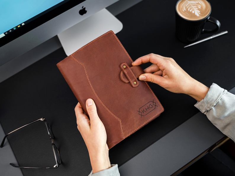 Peut inclure: Un journal en cuir marron avec une fermeture &agrave; boucle. Le journal pr&eacute;sente un motif cousu et les initiales "KHG" sont visibles. Le journal est tenu par une personne. Une tasse de caf&eacute; et un ordinateur sont en arri&egrave;re-plan.