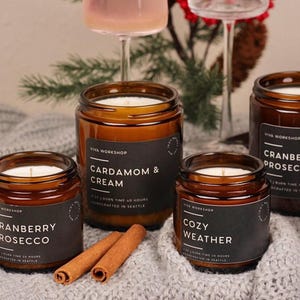 Puede incluir: Cuatro velas en frascos de vidrio &aacute;mbar con etiquetas negras. Las etiquetas muestran el texto Viva Workshop y los nombres de los aromas: Cardamom & Cream, Cozy Weather y Cranberry Prosecco. Cada etiqueta indica 7 oz y 40 horas de combusti&oacute;n.
