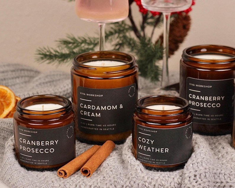 Puede incluir: Cuatro velas en frascos de vidrio &aacute;mbar con etiquetas negras. Las etiquetas muestran el texto Viva Workshop y los nombres de los aromas: Cardamom & Cream, Cozy Weather y Cranberry Prosecco. Cada etiqueta indica 7 oz y 40 horas de combusti&oacute;n.