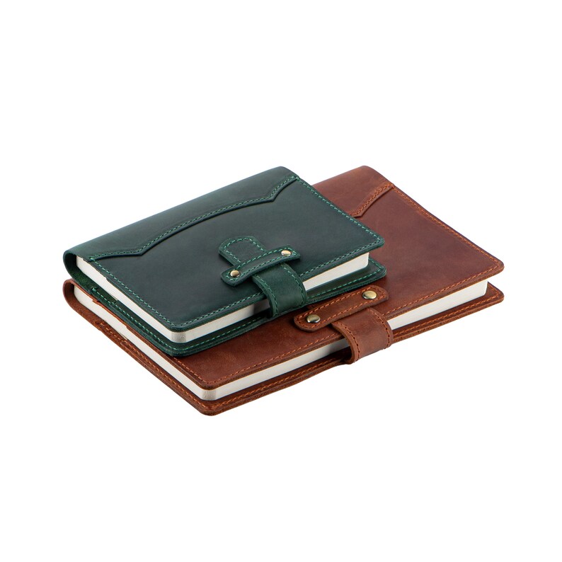 Peut inclure: Deux carnets en cuir, un vert for&ecirc;t et un marron, empil&eacute;s. Le carnet vert a une fermeture &agrave; rabat, le marron une sangle. Les deux ont des int&eacute;rieurs &agrave; pages blanches. Les carnets mesurent environ 15 cm de long.